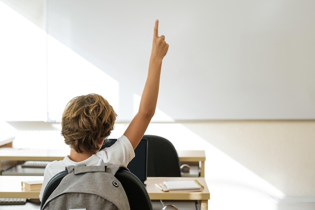 Back view of unrecognizable boy with raised arm sitting at table in classroom during lesson