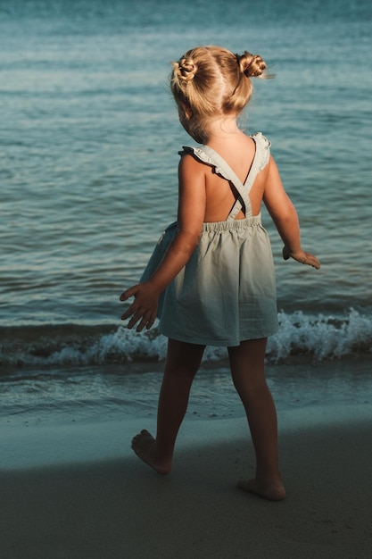 Back view of unrecognizable little girl in blue dress strolling near waving sea in shadow