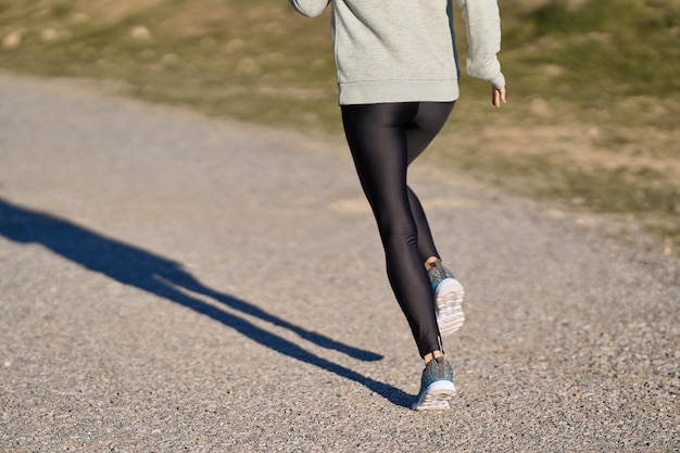 Back view of unrecognizable sportswoman running through park in black leggings on sunny day