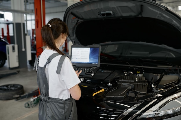 Back view on woman auto engineer doing computer diagnostic using laptop computer for finding car failure causes. Automobile inspection at professional garage service workshop