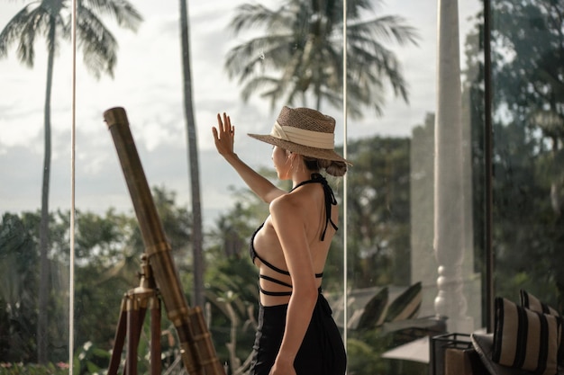 Back view of woman in black dress standing near telescope on a balcony looking at ocean view.