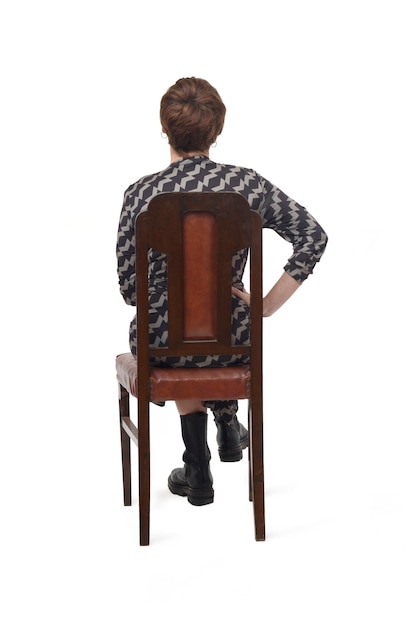 Back view of a woman in dress and boots sitting on chair with croos legged on white background