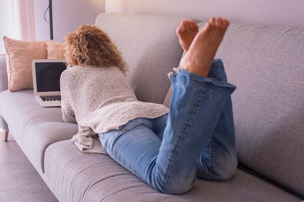 Back view of woman having total relax at home laying dowon on sofa and using laptop to surf the net and enjoy social media online activity One female paople resting on couch in lazy sunday time