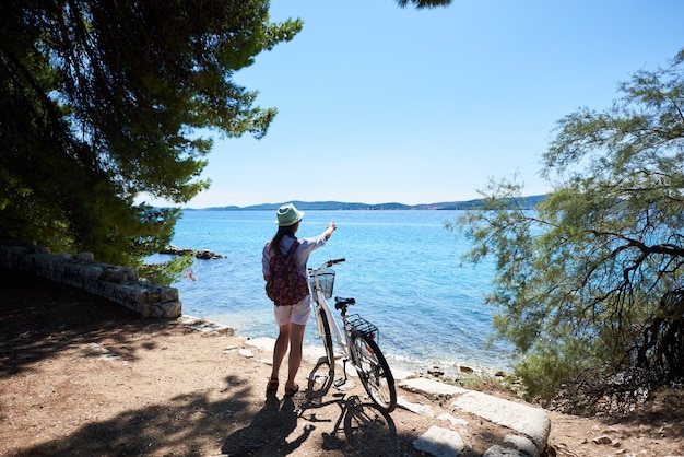 Photo back view of woman standing with bicycle on stony sidewalk under clear blue sky pointing at opposite shore on sparkling clear sea background on summer day.