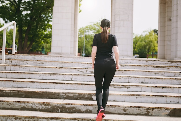 Back view of young athletic strong brunette woman in black uniform and cap doing sport exercises, warm-up before running climbing on stairs in city park outdoors