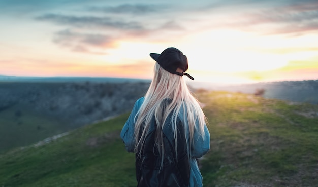 Back view of young blonde girl with black cap and backpack, walking on peak of the hills. Background of sunset.