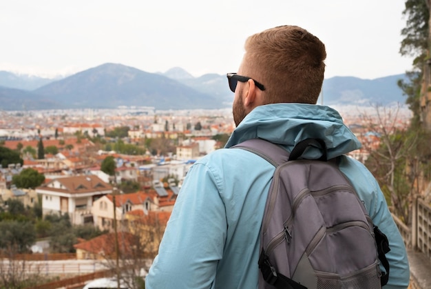 Back view of young caucasian blond bearded young man with backpacktourist looking at beautiful cityred roofs of towncity in mountainsConcept of traveltourismtrip