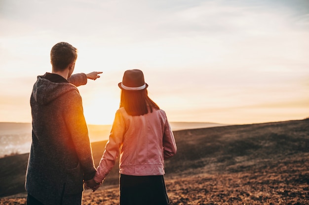 Back view of a young couple traveling around the world while man is show with his finger to his girlfriend where to watch against sunset .