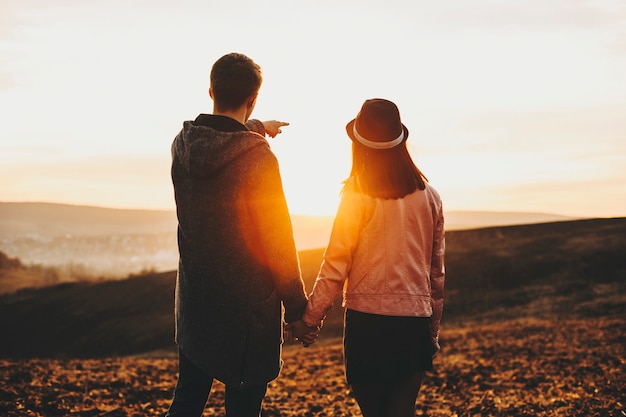 Back view of young guy holding hand of girlfriend and pointing at distance for her while standing in field during beautiful sunset.Unrecognizable couple looking at distance during sundown