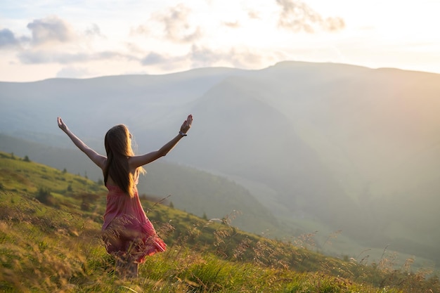 Back view of young happy woman traveler in red dress standing on grassy hillside on a windy evening in summer mountains with outstretched arms enjoying view of nature at sunset