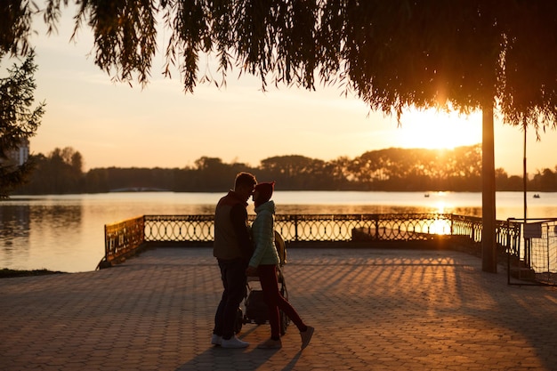 Back view of young loving couple silhouettes holding hands parents with baby pram in autumn park on