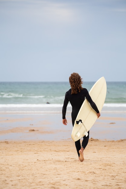 Back view of young man in black wetsuit walking towards sea with surfboard