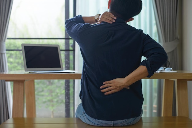 Back view of young man sitting on desk with laptop and suffering from back pain during long hours of work