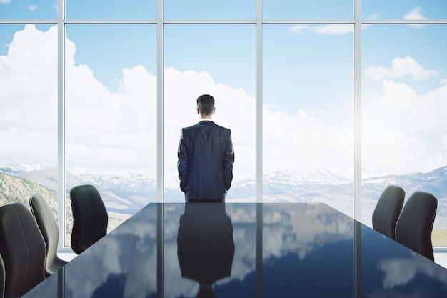 Back view of young man standing in modern meeting room interior with reflections on table chairs and panoramic window with sky clouds view and daylight CEO and conference concept