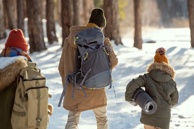 Back view of young man with backpack and little girl with rolled mat moving towards country house, woman with rucksack following them