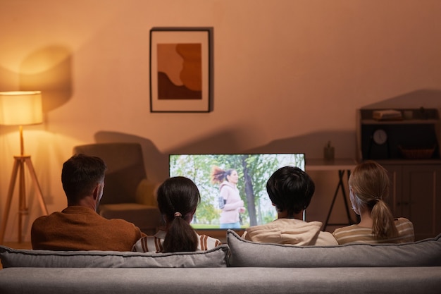 Back view of young people watching movies at home while sitting on sofa, focus on TV screen, copy space