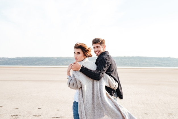 Back view of a young smiling couple hugging and looking at camera at the beach