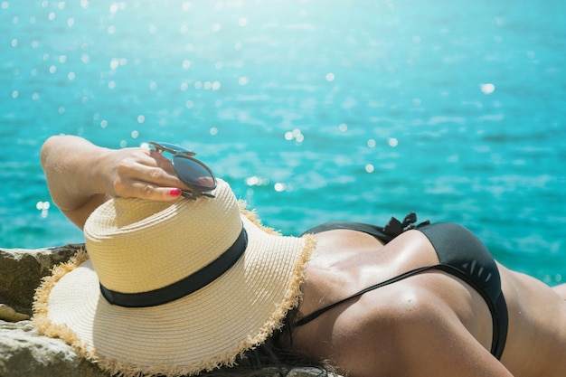 Back view young woman in black swimsuit and straw hat on wild snone beach with clean sea