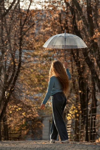 Back view on young woman with long hair walks through autumn park Pretty girl with transparent umbrella