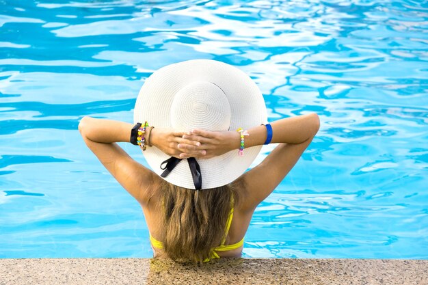 Back view of young woman with long hair wearing yellow straw hat relaxing in warm summer swimming pool with blue water on a sunny day.