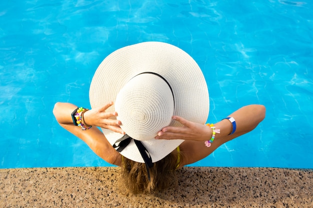 Back view of young woman with long hair wearing yellow straw hat relaxing in warm summer swimming pool with blue water on a sunny day.