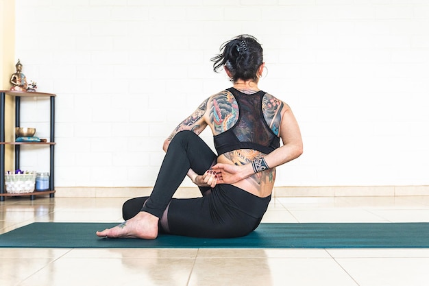 Back woman with tattoos in ardha matsyendrasana yoga pose in front of a white wall with yoga elements