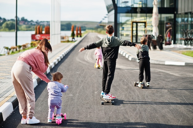 Back of young stylish mother with four kids outdoor. Sports family spend free time outdoors with scooters and skates.