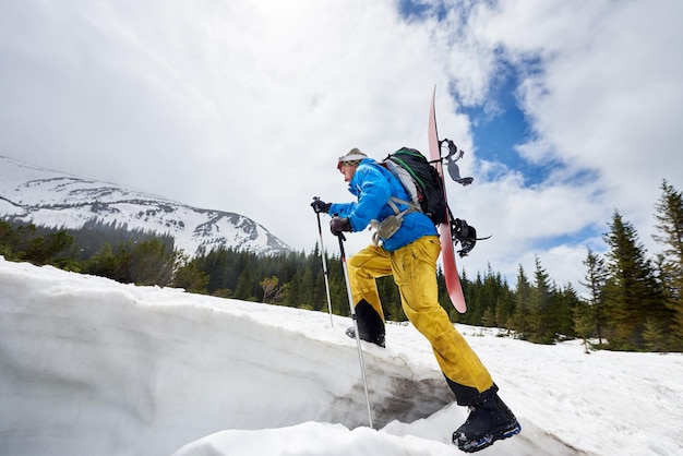 Backcountry skiing Backpacker walking up with snowboard on back Side low angle Sky mountain and woods on background