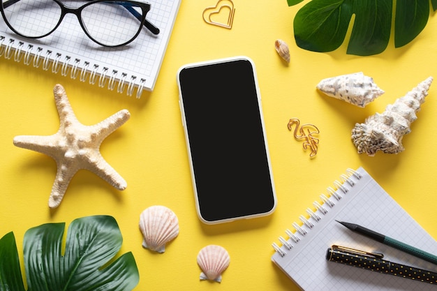 A background with a smartphone glasses a blank notepad and leaves and shells on a yellow background Blogging in the summer at the workplace is a business Copy space Flat lay top view