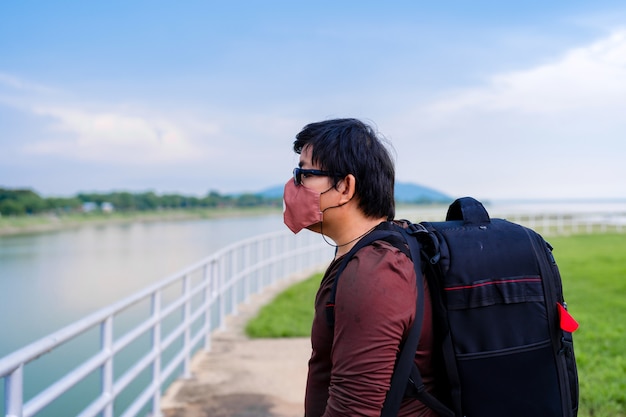 Backpack Asian man wear fabric protective face mask during travel, man relax on vacation standing alone looking at dam lake view