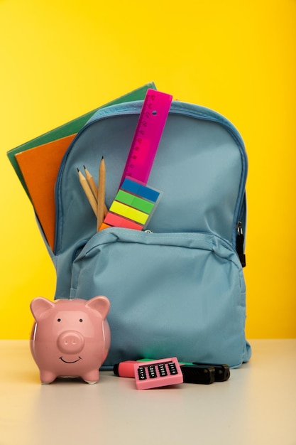 Backpack with school tools and piggy bank on yellow background. Vertical image.