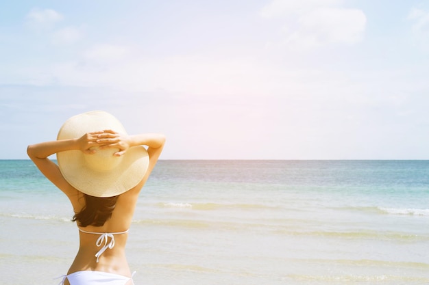 Backside young woman skin tan in sunhat and bikini standing with her arms raised to her head enjoying on the beach vacation travel and enjoy life at sea looking view of beach ocean on hot summer day