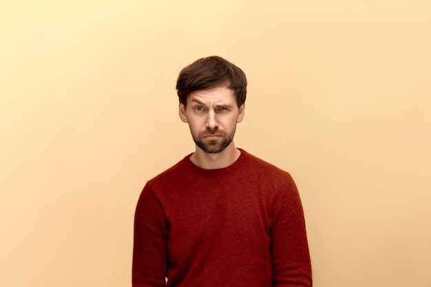 Bad connection. Photo of young man with beard wearing sweater, frowns face, dislikes something, looks with disgusting expression, posing against beige wall.