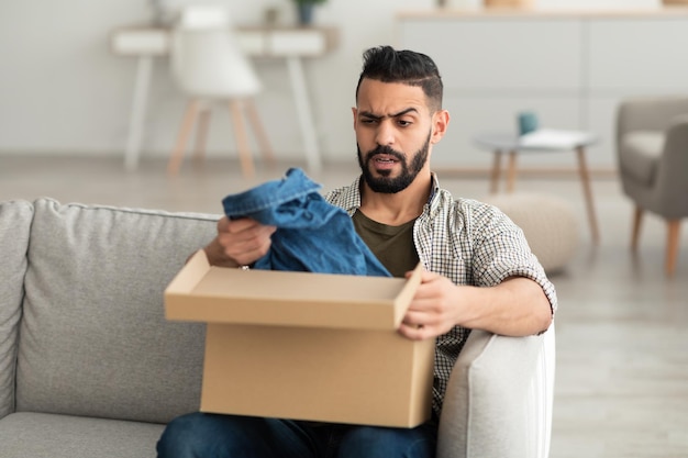 Bad delivery service Angry Arab man unpacking cardboard box receiving wrong item feeling upset with parcel at home