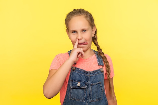 Bad manners Portrait of funny cute little girl with braid in denim overalls picking nose and sticking out tongue having fun child behaving impolite indoor studio shot isolated on yellow background