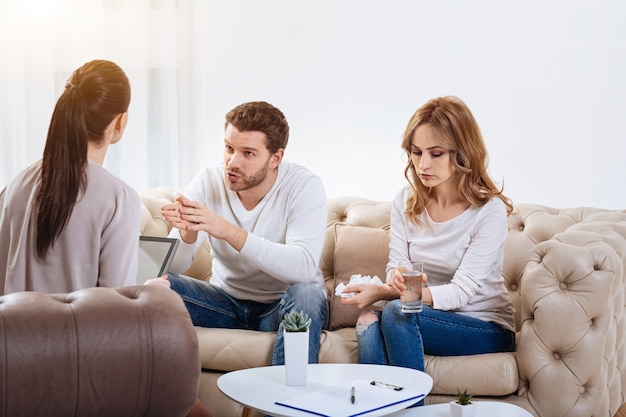 Bad mood. Beautiful unhappy young woman holding a glass of water and a tissue and looking down while listening to her husband