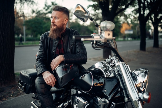 Baerded biker poses on chopper leaning on a helmet. Vintage bike, rider and his motorcycle, freedom lifestyle, biking