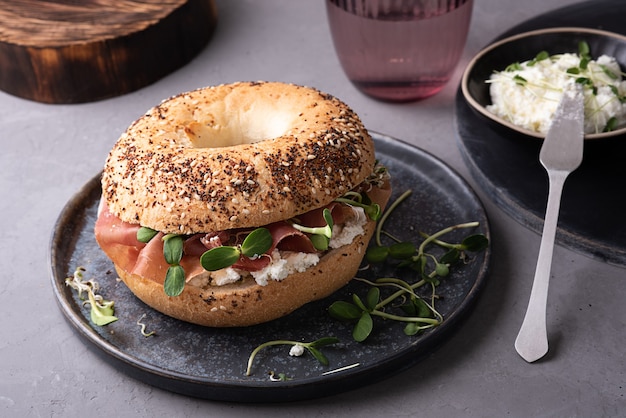 Bagel with cream cheese, dry cured ham and microgreens on a plate on a gray background, breakfast sandwich.