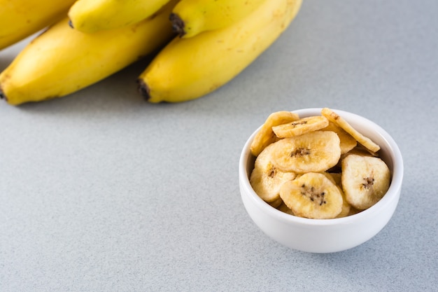 Baked banana chips in a white bowl and a bunch of bananas on the table. Fast food. Close-up. Vertical view. Copy space