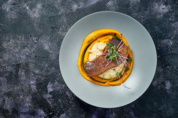 Baked beef tongue with mashed potatoes and carrots in a gray bowl on a black background. Top view. Close up