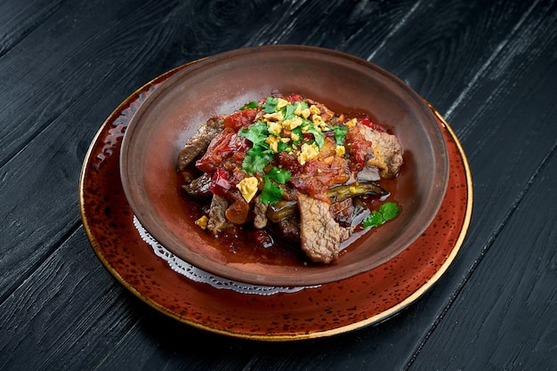 Baked beef with eggplant, nuts, tomatoes and cilantro in clay, served in a bowl on a black background.