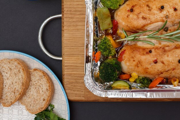 Photo baked chicken breasts or fillet with vegetables and greens in metal container, plate with bread on a wooden cutting board, top view.