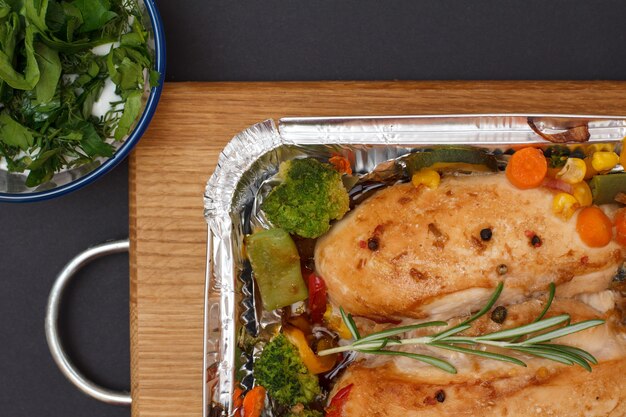 Photo baked chicken breasts or fillet with vegetables and greens in metal container on a wooden cutting board. glass bowl with sauce and rosemary. top view.