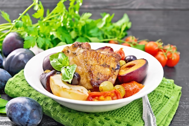 Baked chicken with tomatoes, apples, plums and grapes in a plate on green napkin, garlic, parsley and basil on wooden board background