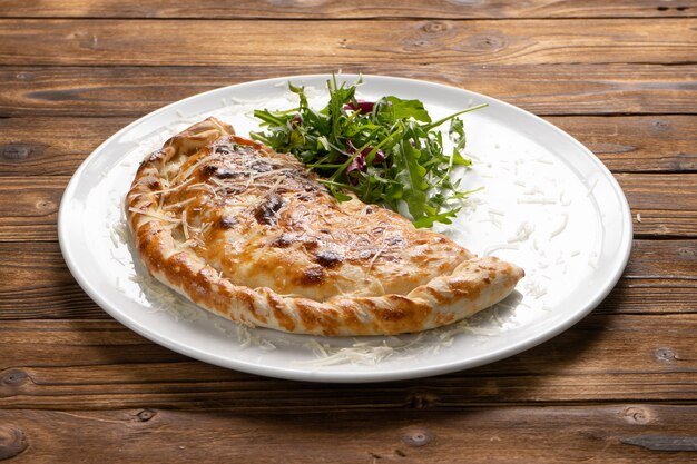 Baked to a crisp brown pizza calzone with fresh arugula and parmesan cheese in a white ceramic plate on a wooden kitchen table.