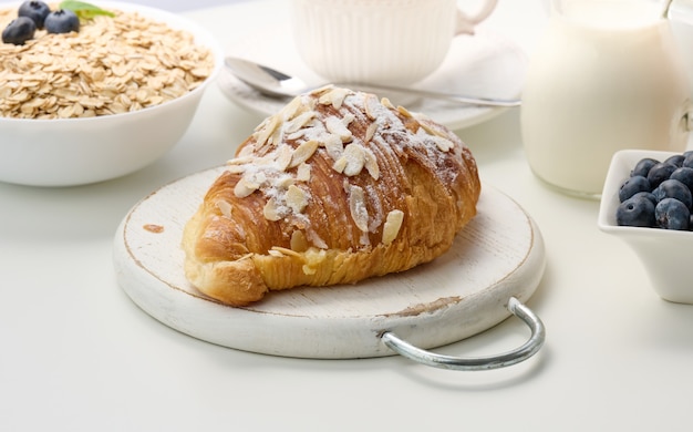 Baked crisp croissant on a wooden board, a plate with blueberry, a white ceramic brew and a cup on a white table. Breakfast