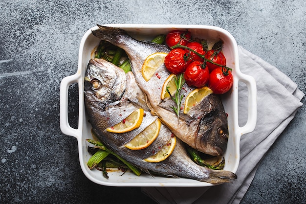 Baked fish dorado with green asparagus and tomatoes in white ceramic baking pan on gray rustic concrete background, top view. Healthy dinner with fish concept, dieting and clean eating