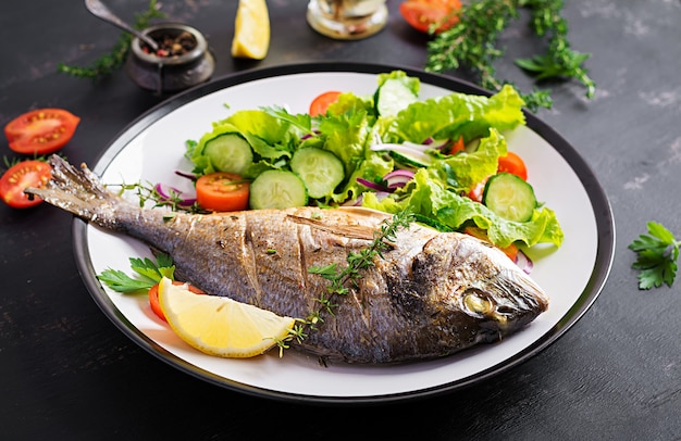 Baked fish dorado with lemon and fresh salad in white plate on dark rustic background. Healthy dinner with fish concept. Dieting and clean eating