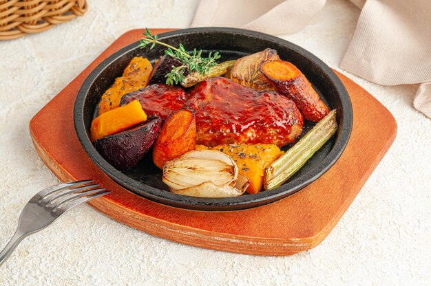 Baked pork in honey sauce. With grilled vegetables and thyme. Served in a frying pan. Nearby is a beige napkin and a fork. Close-up. Light background.