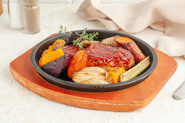 Baked pork in honey sauce. With grilled vegetables and thyme. Served in a frying pan. Nearby is a beige napkin and spices. Close-up. Light background.